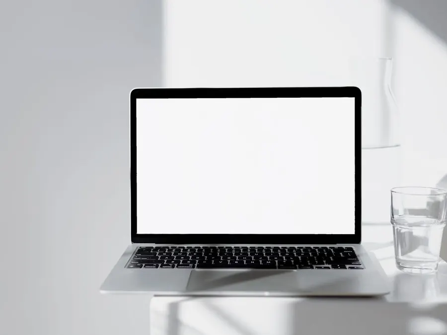 A glass of water standing next to an open laptop on a white desk.
