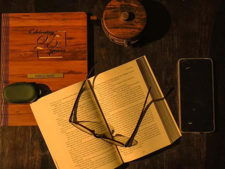 An open book with reading glasses resting on it and a dark phone lying beside it, on a wooden table.